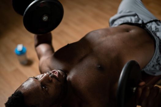 Shirtless afro american man doing exercises with dumbbells lying on bench in gym