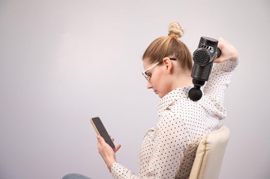 A young woman sits on a chair, uses a smartphone and makes herself massage with an electro massage gun