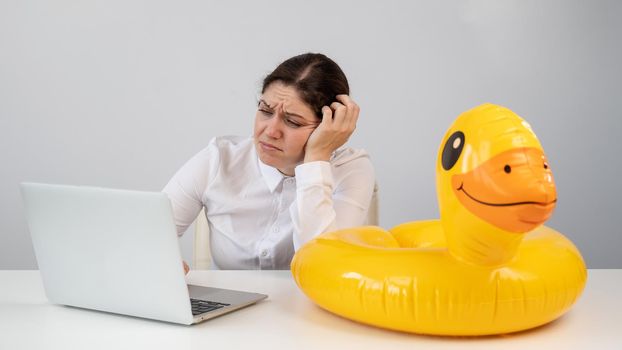 Caucasian woman sits at a table with a laptop and an inflatable duck on a white background. Office worker dreaming of vacation