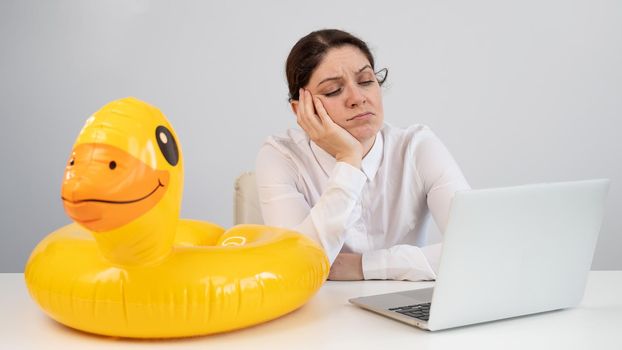 Caucasian woman sits at a table with a laptop and an inflatable duck on a white background. Office worker dreaming of vacation