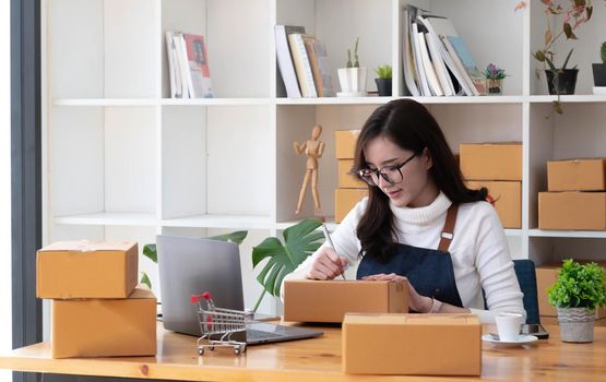 Young beautiful happy asian business woman owner of SME online using laptop receive order from customer with parcel box packaging at her startup home office, online business seller and delivery.