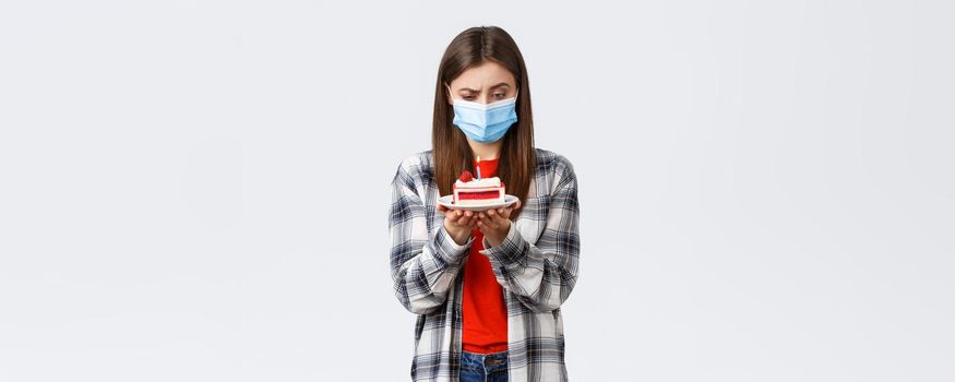 Coronavirus outbreak, lifestyle during social distancing and holidays celebration concept. Confused woman in medical mask staring puzzled at birthday cake lit candle, thinking, white background.