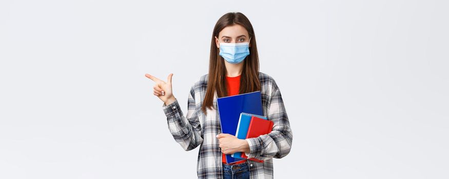 Coronavirus pandemic, covid-19 education, and back to school concept. Young pretty female student in medical mask with notebooks, pointing finger left, showing university info.