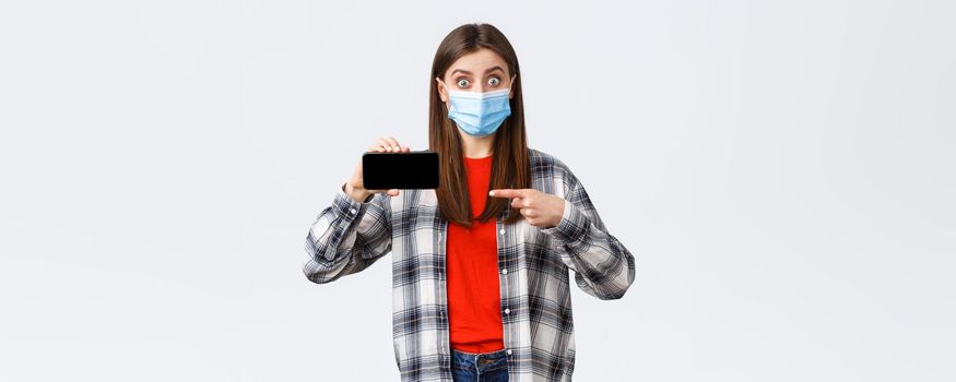 Different emotions, covid-19, social distancing and technology concept. Impressed and surprised girl in medical mask stare astonished, pointing finger at mobile phone screen.