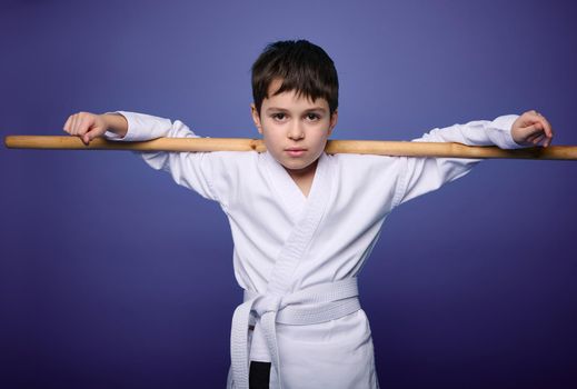 Confident charming European aikido wrestler posing with wooden weapon against violet background with copy ad space for advertising text. Oriental martial arts practice
