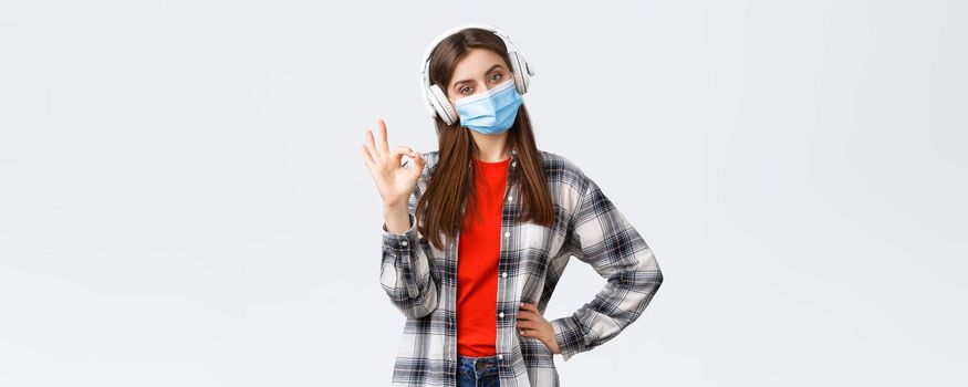 Social distancing, leisure and lifestyle on covid-19 outbreak, coronavirus concept. Pleased good-looking woman in medical mask and headphones, listening music, show okay sign, approve or like.