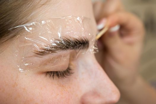 The master applies the composition for laminating the eyebrows to the client with a wooden spatula