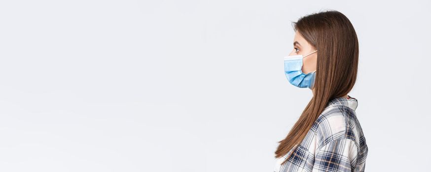 Coronavirus outbreak, leisure on quarantine, social distancing and emotions concept. Profile of serious-looking young pretty woman in medical mask standing in line, white background.