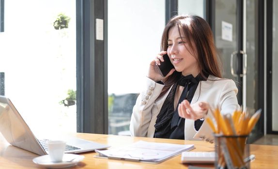 Happy young Asian businesswoman using smartphone and laptop computer at office..