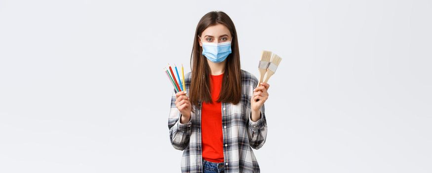 Social distancing, leisure and hobbies on covid-19 outbreak, coronavirus concept. Good-looking pleased girl in medical mask showing painting brushes and pencils, recommend start drawing.