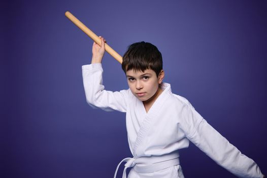 Confident charming European aikido wrestler poses with wooden weapon during Oriental martial arts practice, isolated on violet background, copy ad space for text. Oriental martial arts practice