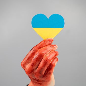 Woman with hands covered in blood holding a heart with the flag of ukraine on a white background