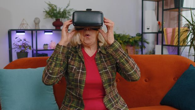 Senior elderly woman using headset helmet app to play simulation game, simulation glasses gadget technology. Watching virtual reality 3D 360 video. Mature grandmother in VR goggles on couch at home