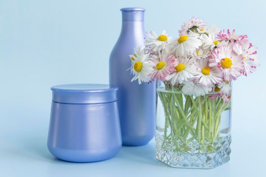 A bouquet of fresh flowers, daisies on green stems are in a glass with clean water and two purple jars with cream cosmetics on a blue background