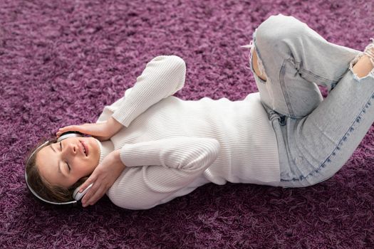 Teenage girl in braids lies on purple carpet with wireless headphones and listens to music. A gentle girlish smile.