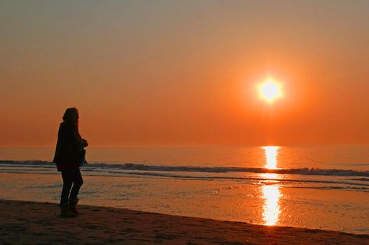 Silhouette of a woman looking at the golden sun sinking in  the ocean. The sky is colored orange. Location: Noordwijk aan Zee, Netherlands