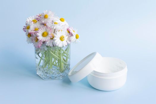 A bouquet of fresh flowers, daisies on green stems are in a glass with clean water and a white jar of cream on a blue background