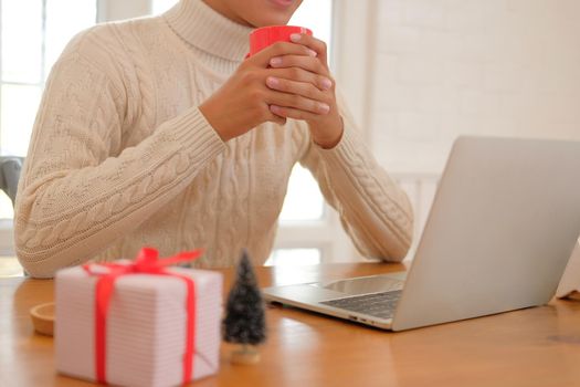 man wearing cream sweater holding coffee cup & using computer at home during christmas. xmas new year celebration