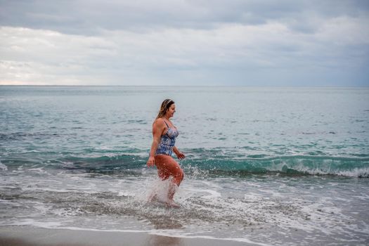 A plump woman in a bathing suit enters the water during the surf. Alone on the beach, Gray sky in the clouds, swimming in winter