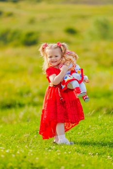 girl in a red dress and with a doll in her hands stands in nature