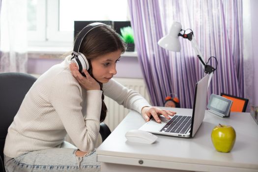 A student is studying remotely using the internet and a laptop. The young girl has wireless headphones worn over her ears.