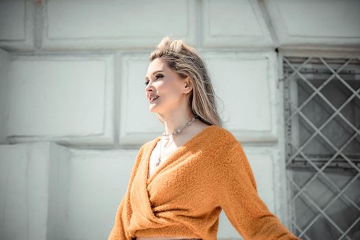 A middle-aged woman looks like a good blonde with curly beautiful hair and makeup on the background of the building. She is wearing a yellow sweater