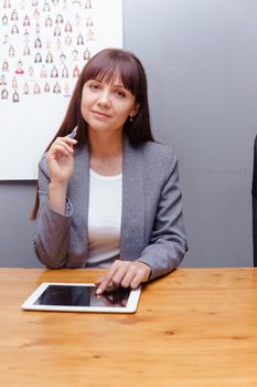 A brunette businesswoman in a gray jacket at her desk with a tablet in her hands. Business portrait in the office.