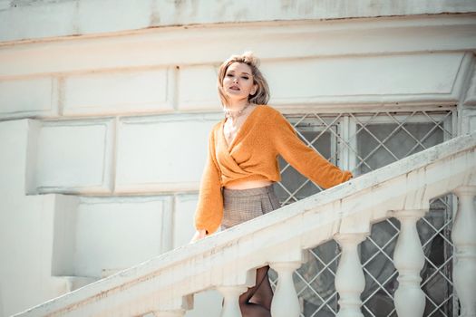 A middle-aged woman looks like a good blonde with curly beautiful hair and makeup on the background of the building. She is wearing a yellow sweater