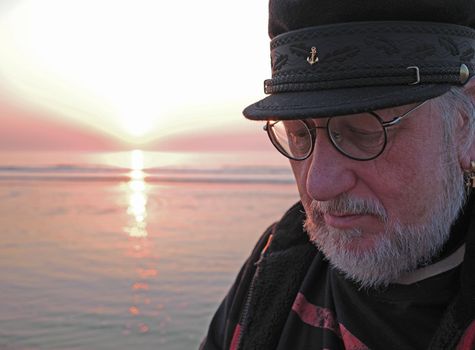 An elderly man is looking thoughtfully down. He is standing in front of the sea. The sun is going down at the horizon. The man has a white beard and a sailor cap on his head
