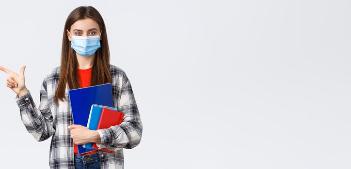 Coronavirus pandemic, covid-19 education, and back to school concept. Young pretty female student in medical mask with notebooks, pointing finger left, showing university info.