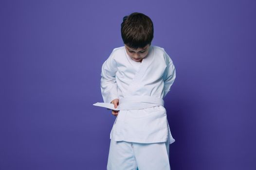 Horizontal portrait of a handsome confident European school age child boy putting on a white kimono, tying a belt around his waist, ready for combat sports. Aikido fighter. Oriental martial arts