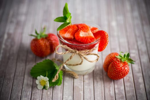 sweet homemade yogurt with strawberry jam and fresh strawberries in a glass cup, on a wooden table.