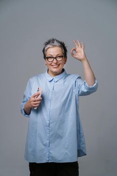 Gesturing OK sign grey haired mature businesswoman with smartphone in hand looking at camera wearing blue shirt. Pretty woman in blue shirt isolated on white. Toned image.