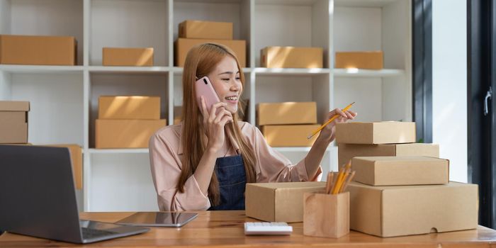 Young Asian small business owner working at home office, taking note on purchase orders. Online marketing packaging delivery, startup SME entrepreneur or freelance woman concept.