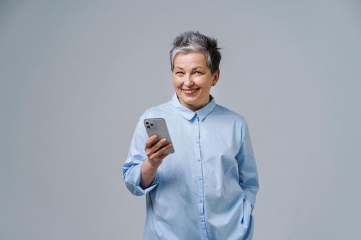 Mature grey haired businesswoman with smartphone in hand looking at camera shopping online, checking social media. Pretty woman in blue shirt isolated on white background. Copy space.