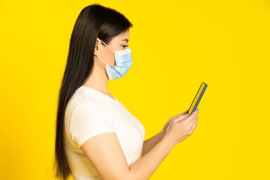 Half length profile of asian middle aged woman in medical face mask, holding smartphone in hands reading news or important information.