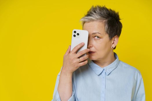 Grey haired mature woman hide face behind smartphone posting negative, toxic comment online, social media. Pretty woman with phone in blue shirt isolated on yellow background.