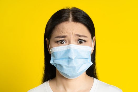 Scared of pandemic mature asian woman wearing medical face mask coronavirus or monkeypox prevention. Charming middle age woman in white t-shirt and medical mask on yellow background.