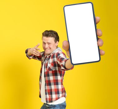 Happy to win handsome man pointing at smartphone with white empty screen, wearing red plaid shirt and jeans cellphone display mock up isolated on yellow background. Mobile app advertisement.