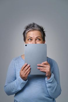 Grey haired mature woman scared hide face behind digital tablet posting negative, toxic comment online, social media. Pretty woman with tablet in blue shirt isolated on white background.