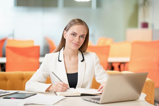 Young business woman in white jacket or suit at the desk with a laptop, looking at camera and making notes. Blonde model female with blue eyes posing in business suit in the office. Business concept.