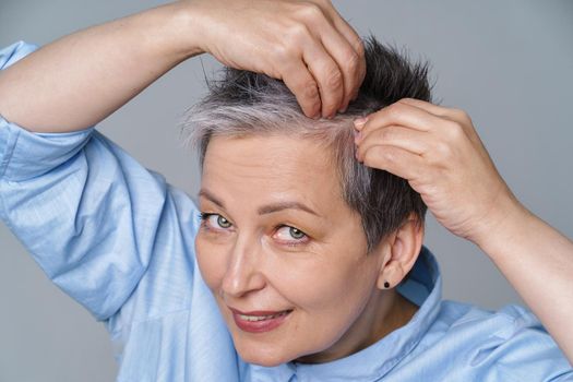 Pretty woman in 50s checking her hair or skull skin in mirror. Beautiful grey haired woman dealing with dandruff problem checking in mirror. Healthy hair, hair loss concept.