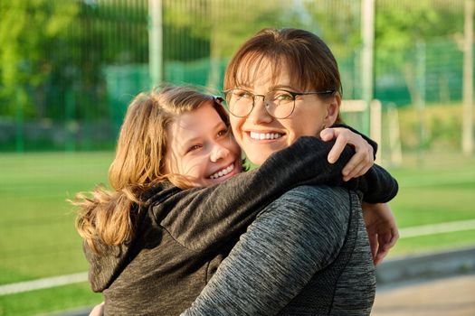 Portrait of happy mom and preteen daughter hugging together outdoor, near sports stadium, football field. Family, happiness, leisure, lifestyle, relationship, love, mother's day, motherhood concept