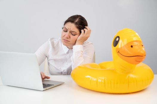 Caucasian woman sits at a table with a laptop and an inflatable duck on a white background. Office worker dreaming of vacation