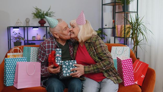 Happy old senior elderly family couple grandparents man woman celebrating birthday anniversary on couch at home. Mature grandmother wife giving gift box surprise present to her husband grandfather