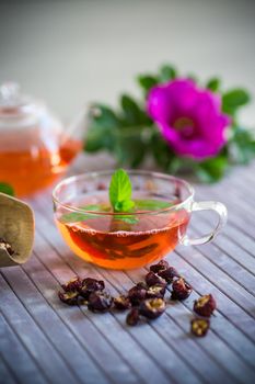 brewed rosehip tea in a glass teapot with rosehip flowers and mint, on a wooden table.