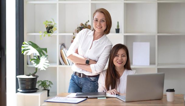 Two middle-aged women are looking at the camera while sitting on a desk in the office. female executives working in the office discussing documents.