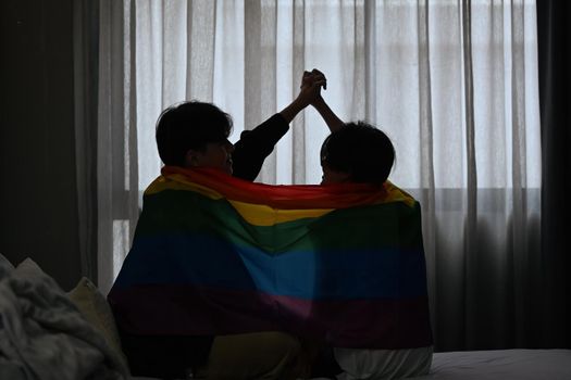 Silhouette of gay couple expressing their love to each other under pride flag. LGBT, pride, relationships and equality concept.