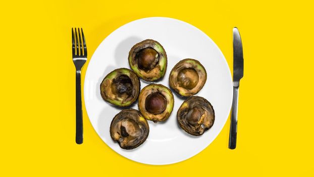 Bad Avocado halfs placed on plate with fork and knife Yellow background. Rotten Food Contaminated Avocado Necomestibile Fruits Wasting Food Unhealthy Tropical Fruits. Food concept Copy space.
