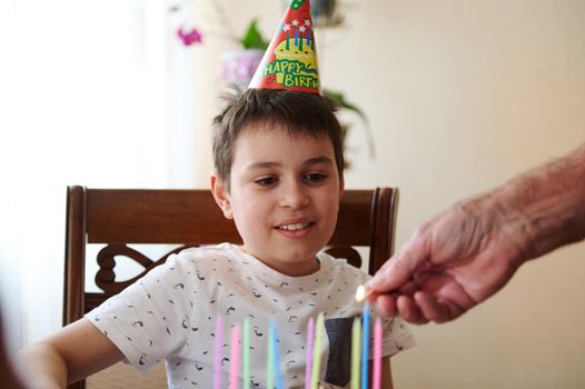 Adorable Caucasian teenage boy looking at grandfather's hand lighting colorful candles on his birthday cake